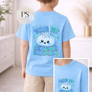 May include: A light blue t-shirt with a cartoon dumpling design and the text "YOUR MY SQUASHIE DUMPLING". The shirt is worn by a child. The design includes blue stars and a cute dumpling illustration.