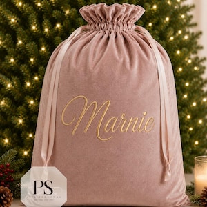 May include: A dusty rose velvet drawstring gift bag featuring the name Marnie embroidered in gold cursive script on the front. The bag has a ruffled top edge and matching satin ribbon ties that cinch the opening closed for a secure closure.