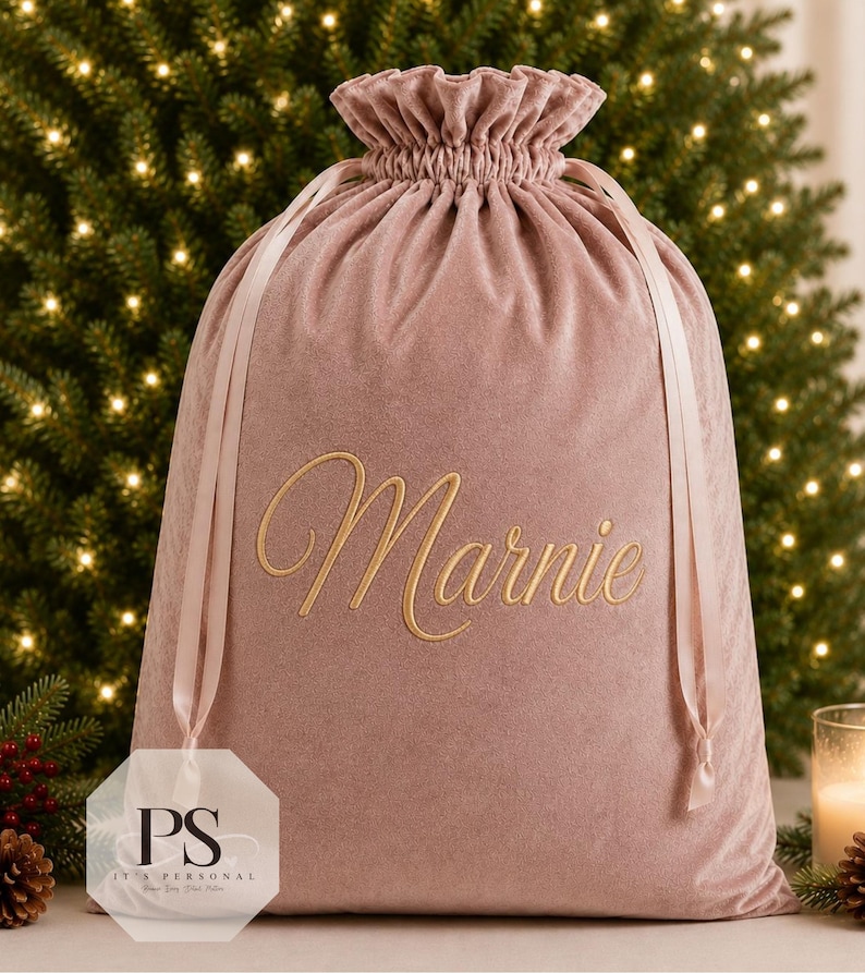 May include: A dusty rose velvet drawstring gift bag featuring the name Marnie embroidered in gold cursive script on the front. The bag has a ruffled top edge and matching satin ribbon ties that cinch the opening closed for a secure closure.