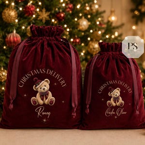 May include: Two deep red velvet drawstring bags featuring a graphic of a teddy bear wearing a hat and scarf. The text CHRISTMAS DELIVERY is printed above the bear, with the names Romey and Caiden Oliver printed below in a script font.