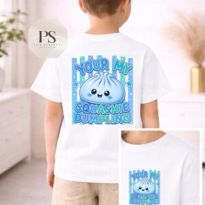 May include: White t-shirt with a blue dumpling graphic and the text "YOUR MY SQUASHIE DUMPLING". The design includes blue stripes and stars. The shirt is worn by a child.