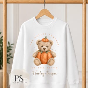 May include: A white long-sleeve sweatshirt featuring a graphic of a light brown teddy bear sitting inside a large orange pumpkin with a smaller pumpkin on its head. The text reads THE CUTEST PUMPKIN IN THE PATCH and the name Harley-Regan.