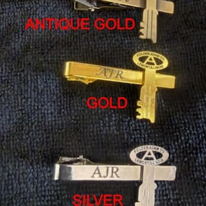 Puede incluir: Tres pasadores de corbata metálicos en oro antiguo, oro y plata. El pasador de oro antiguo está grabado con "Root", el pasador de oro con "AJR" y el pasador de plata con "AJR". Cada pasador presenta un diseño en forma de llave con el texto "FOLDER ADAM CO LEMONT ILLINOIS".