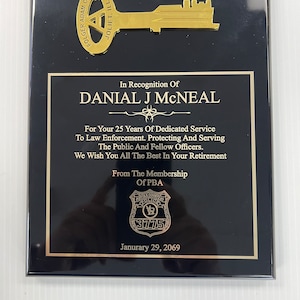 May include: A black plaque with gold lettering and a gold key design. The plaque reads "In Recognition Of Danial J McNeal For Your 25 Years Of Dedicated Service To Law Enforcement. Protecting And Serving The Public And Fellow Officers. We Wish You All The Best In Your Retirement From The Membership Of PBA Folk County Police 3775 Janurary 29, 2069"