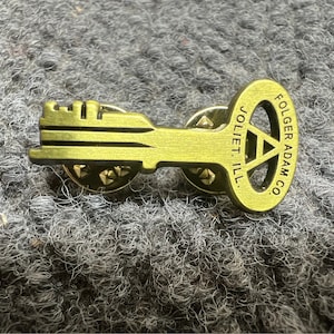 May include: A gold-toned key-shaped pin with the text "FOLGER ADAM CO. JOLIET ILL." on the circular part of the key.