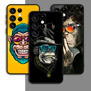 Op de afbeelding: Drie telefoonhoesjes met verschillende apenontwerpen. De eerste hoes heeft een blauwe aap met een zonnebril op een gele achtergrond. De tweede hoes heeft een zwart-witte aap met een hoed en zonnebril op een zwarte achtergrond. De derde hoes heeft een bruine aap met een zonnebril op een zwarte achtergrond.