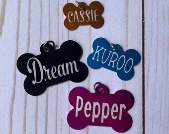 Puede incluir: Cuatro etiquetas para mascotas en forma de hueso en bronce, negro, azul y magenta. Cada etiqueta tiene un nombre diferente en escritura blanca: "Cassie", "Dream", "Kuroo" y "Pepper". Todas las etiquetas tienen un pequeño anillo de metal para sujetar.