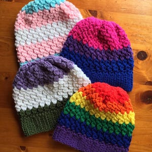 Op de afbeelding: Vier kleurrijke gehaakte mutsen worden getoond op een houten oppervlak. De mutsen hebben verschillende kleurencombinaties, waaronder roze, witte en blauwe strepen; roze, paarse en blauwe verlopen; paarse, witte en groene strepen; en een regenboogpatroon.