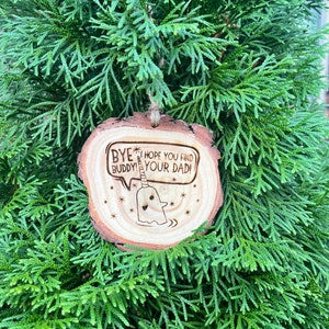 Op de afbeelding: Ronde houten ornament met een narwal illustratie en de tekst "Bye Buddy! Hope You Find Your Dad!". Het ornament hangt aan een natuurlijke touwtje en staat tegen een achtergrond van groen gebladerte.