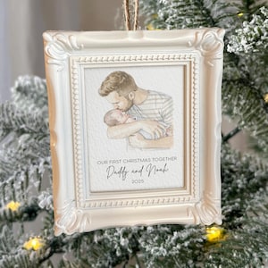 May include: A white framed Christmas ornament with a watercolor image of a father kissing a baby. The text reads "OUR FIRST CHRISTMAS TOGETHER Daddy and Noah 2025". The ornament hangs from a brown string.