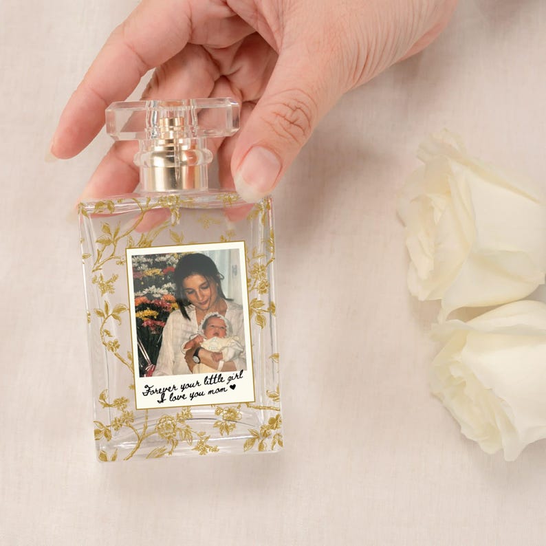 May include: Clear rectangular perfume bottle with a gold floral design and a photo of a mother and child. The bottle has a clear, rectangular cap and gold accents. The text on the photo reads "Forever your little girl I love you mom."