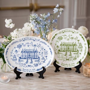 May include: Two oval ceramic plates on black stands. The left plate features blue ink illustrations of a chateau, wedding icons, and the text David & Jenna, November 02, 2026, Chateau du Grand-Luck. The right plate has green ink with Kiera & James, November 09, 2026, Chateau de Tourreau.