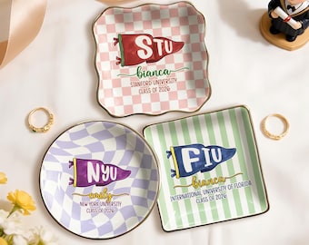 May include: Three decorative ceramic trays with gold trim. One is square with pink and white checkered pattern and a red pennant that says "STU". Another is round with a purple and white checkered pattern and a purple pennant that says "NYU". The third is square with green stripes and a blue pennant that says "FIU".