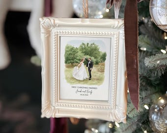 May include: A white framed ornament with a watercolor painting of a couple dancing in a field. The text "First Christmas Married" and names are below the image. The ornament is hanging on a Christmas tree with other ornaments and a brown ribbon.
