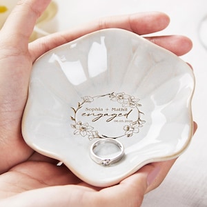 May include: A white, petal-shaped ceramic ring dish with a silver ring. The dish has a floral wreath design with the words "Sophia + Mathil engaged 06.05.2026" in brown script.