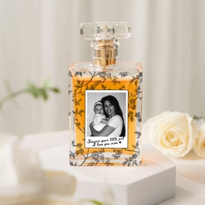 May include: A clear perfume bottle with a gold-colored liquid inside. The bottle has a clear, faceted stopper and a gold-colored neck. A black and white photo of a mother and child is on the front, with the text "Forever your little girl I love you mom."