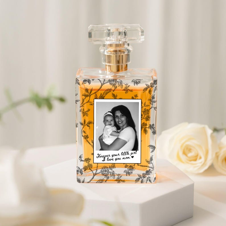 May include: A clear perfume bottle with a gold-colored liquid inside. The bottle has a clear, faceted stopper and a gold-colored neck. A black and white photo of a mother and child is on the front, with the text "Forever your little girl I love you mom."