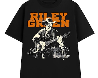 May include: Black t-shirt featuring a graphic of a musician playing a guitar, with the text "RILEY GREEN" in orange block letters above the image. The image is a sepia-toned illustration of the musician. The shirt is a classic crew neck style.