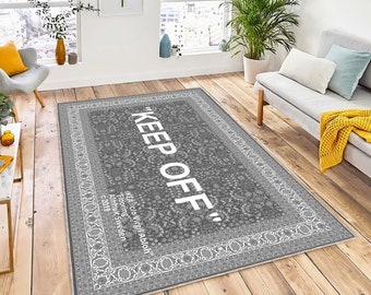 Könnte beinhalten: Ein grauer und weißer Teppich mit Muster und dem Text "KEEP OFF" in weißen Buchstaben. Der Text ist von einem weißen Rand mit floralem Muster umgeben. Der Teppich liegt auf einem Holzboden.