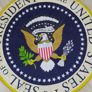 Puede incluir: Una alfombra redonda azul y dorada con el Sello Presidencial de los Estados Unidos. El sello presenta un &aacute;guila calva con un escudo, una rama de olivo y flechas. El texto "Sello del Presidente de los Estados Unidos" est&aacute; escrito alrededor del sello.