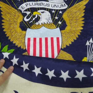 Puede incluir: Un primer plano de una tela azul y blanca con un borde amarillo. La tela presenta un gran &aacute;guila blanca con rayas rojas, blancas y azules en el pecho. El &aacute;guila sostiene un rollo con las palabras "E Pluribus Unum" en amarillo. La tela est&aacute; rodeada por un anillo de estrellas blancas sobre un fondo azul.