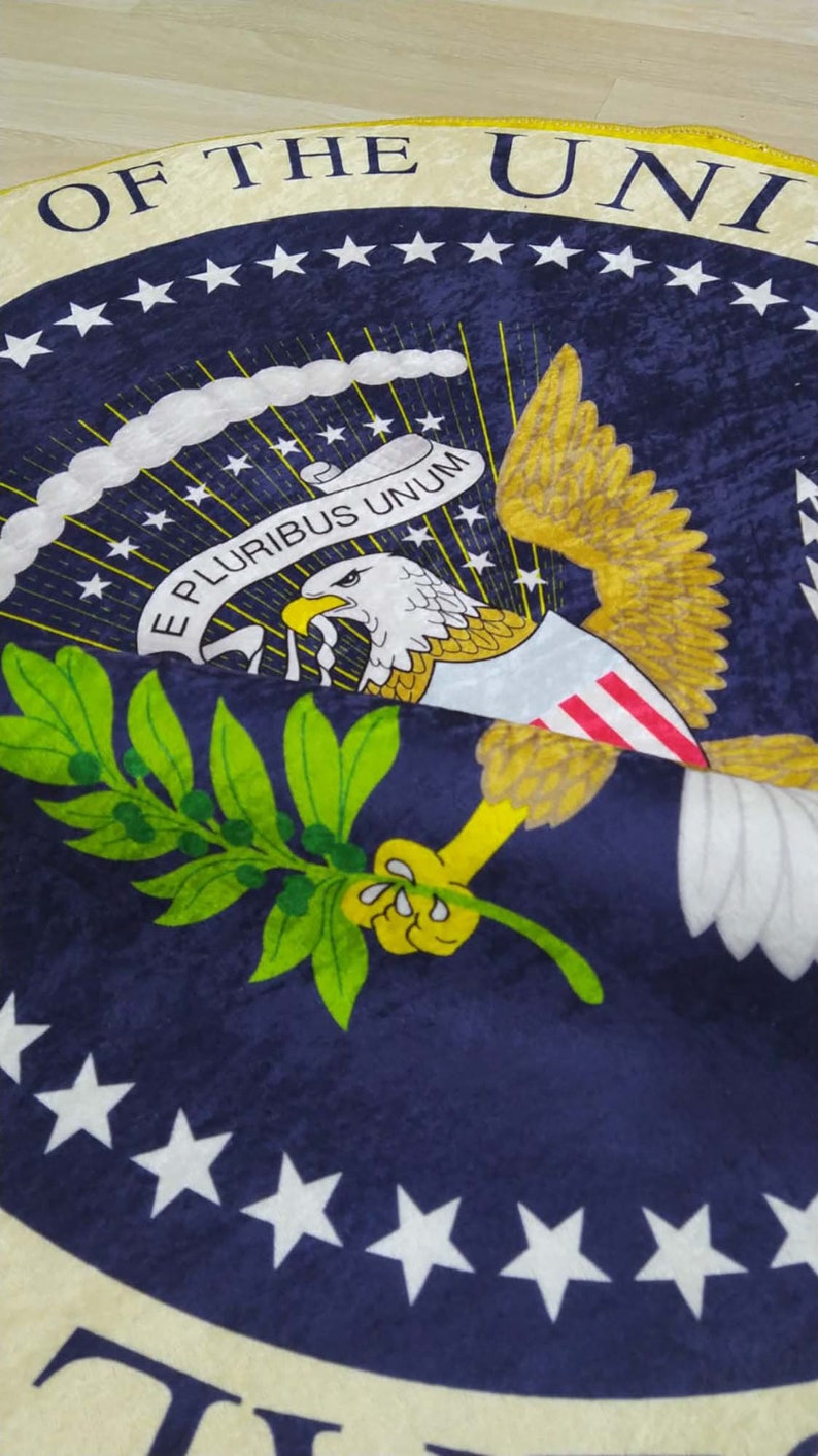 Puede incluir: Una alfombra circular azul y dorada con el Gran Sello de los Estados Unidos. El sello representa un &aacute;guila calva con las alas extendidas, sosteniendo una rama de olivo en su garra derecha y flechas en su garra izquierda. El &aacute;guila est&aacute; posada sobre un escudo con trece rayas y un campo azul con trece estrellas. El sello est&aacute; rodeado por un anillo de estrellas blancas y las palabras "E Pluribus Unum" y "Of the United States".