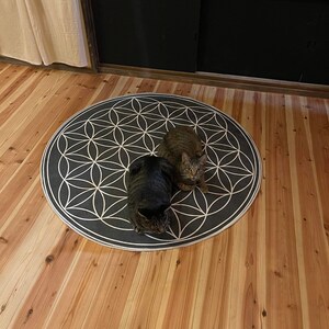 May include: Two cats, one brown tabby and one black cat, are lying on a gray and white circular rug with a flower of life pattern. The rug is on a wooden floor.