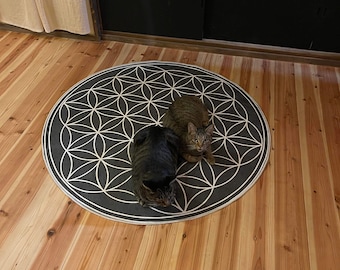 Op de afbeelding: Twee katten, een bruine tabby en een zwarte kat, liggen op een grijs en wit rond tapijt met een levensbloem-patroon. Het tapijt ligt op een houten vloer.
