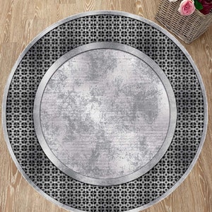 May include: A round, silver-colored rug with a black and white geometric pattern around the edge. The center of the rug is a solid silver color with a distressed, aged look.