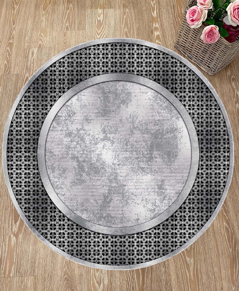 May include: A round, silver-colored rug with a black and white geometric pattern around the edge. The center of the rug is a solid silver color with a distressed, aged look.
