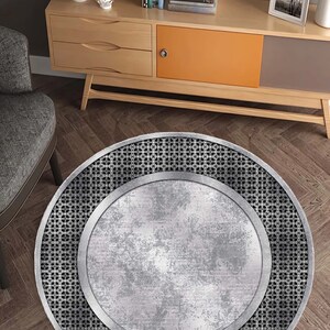 May include: A round, silver-colored rug with a black, geometric pattern around the edge. The rug has a distressed, metallic look.