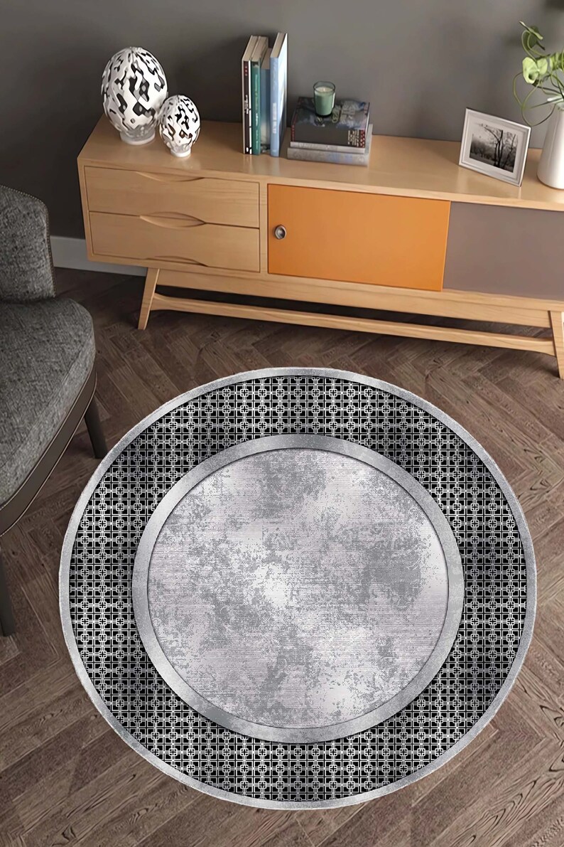 May include: A round, silver-colored rug with a black, geometric pattern around the edge. The rug has a distressed, metallic look.