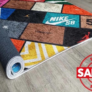 Puede incluir: Una alfombra colorida con un patr&oacute;n geom&eacute;trico que presenta varios logotipos de marcas, incluyendo Nike, SB y 500. La alfombra tiene un borde blanco y est&aacute; enrollada.