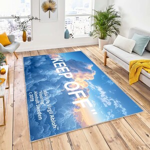 Op de afbeelding: Een blauw en wit tapijt met een wolkenpatroon en de tekst "KEEP OFF" in wit. De tekst "IKEA c/o Virgil Abloh" is ook inbegrepen, samen met "Flooring", "Almhult Sweden" en "c 2019".