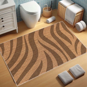 May include: A rectangular bathroom mat with a zebra stripe pattern in shades of brown and tan. The mat has a textured surface and is placed on a wooden floor. The design features wavy stripes, creating a natural look for the bathroom.