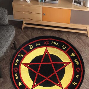 Puede incluir: Una alfombra redonda negra y amarilla con un pentagrama rojo en el centro. La alfombra tiene un borde rojo con texto negro alrededor del borde. El texto incluye las palabras "AL", "PHA", "OME" y "GA".
