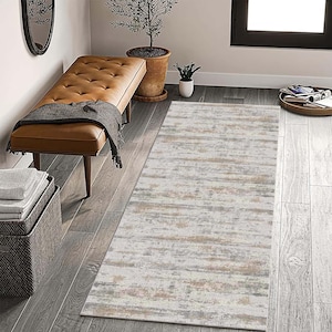 May include: A rectangular area rug with a distressed, abstract design. The rug features a blend of neutral colors, including off-white, gray, and tan, creating a textured, weathered appearance. The rug is placed on a wooden floor.