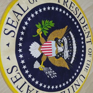 Puede incluir: Una alfombra redonda con fondo azul y borde amarillo. El centro de la alfombra presenta el Sello del Presidente de los Estados Unidos, que incluye un &aacute;guila calva con un escudo, ramas de olivo y flechas. El texto "SEAL OF THE PRESIDENT OF THE UNITED STATES" est&aacute; escrito alrededor del borde exterior del sello.
