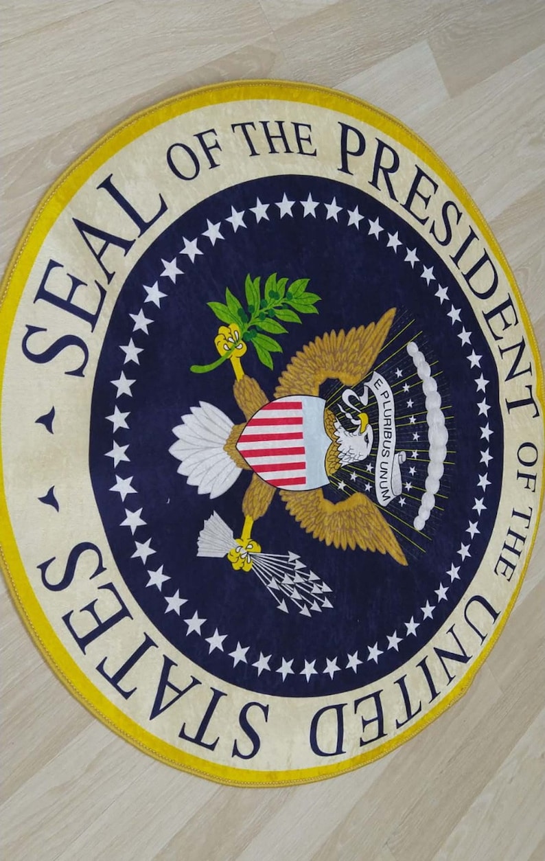 Puede incluir: Una alfombra redonda con fondo azul y borde amarillo. El centro de la alfombra presenta el Sello del Presidente de los Estados Unidos, que incluye un &aacute;guila calva con un escudo, ramas de olivo y flechas. El texto "SEAL OF THE PRESIDENT OF THE UNITED STATES" est&aacute; escrito alrededor del borde exterior del sello.