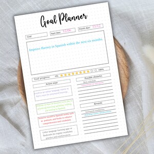 Puede incluir: Un planificador de objetivos imprimible con un fondo punteado. El planificador se titula "Goal Planner" e incluye secciones para el objetivo, la fecha de inicio, la fecha de finalizaci&oacute;n, el progreso del objetivo, los pasos a seguir, los posibles obst&aacute;culos y las recompensas. El objetivo es "Mejorar la fluidez en espa&ntilde;ol dentro de los pr&oacute;ximos seis meses."