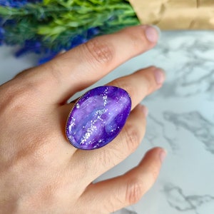 May include: A purple and white galaxy-themed ring with silver glitter accents. The ring is oval shaped and has a gold band.
