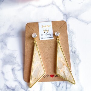 Op de afbeelding: Een paar gouden driehoekige oorbellen met witte en gouden marmeraccenten en heldere kristallen. De oorbellen worden getoond op een bruine kaart met de tekst "'Evótion' Clay Jewelry @enation.clay.jewelry made with ❤️"
