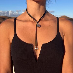 Puede incluir: Un collar lariat de cuero negro con un colgante esf&eacute;rico plateado y dos esferas plateadas m&aacute;s peque&ntilde;as al final de los cordones colgantes. El collar se lleva sobre un top negro.