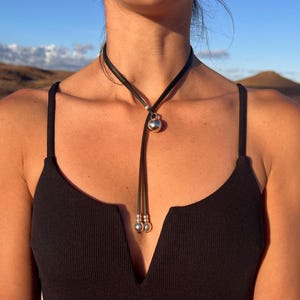 Puede incluir: Un collar lariat de cuero negro con un colgante esf&eacute;rico plateado y dos esferas m&aacute;s peque&ntilde;as en los extremos. El collar se lleva sobre un top negro, con un fondo natural.