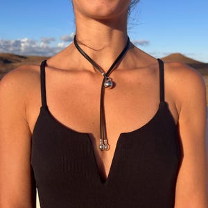 Puede incluir: Un collar lariat de cuero negro con un colgante en forma de coraz&oacute;n plateado y dos peque&ntilde;os detalles de bolas plateadas. El collar se lleva sobre un top negro, con un fondo natural al aire libre.