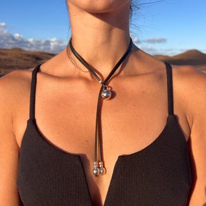 Puede incluir: Collar gargantilla de cuero negro con un colgante esf&eacute;rico plateado y dos cuentas plateadas m&aacute;s peque&ntilde;as. El collar se lleva alrededor del cuello. El fondo es una escena borrosa al aire libre.
