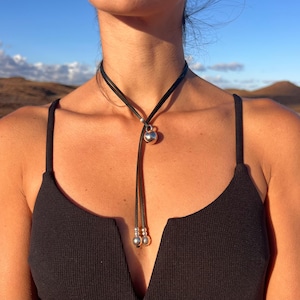 Puede incluir: Un collar gargantilla de cuero negro con un colgante esf&eacute;rico plateado y dos cuentas plateadas m&aacute;s peque&ntilde;as al final de los cordones colgantes. El collar se lleva sobre un top negro.