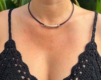 May include: A black leather choker necklace with a silver-colored curved bar pendant. The necklace is worn against a person's skin, with a black crochet top visible in the foreground. Green foliage is in the background.
