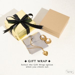 Pu&ograve; includere: Una collana d'oro con un ciondolo a forma di cuore e un paio di orecchini a cerchio d'oro sono esposti su una pochette di velluto grigio. I gioielli sono confezionati in una scatola regalo dorata con un nastro nero e una scatola regalo marrone. Il testo "GIFT WRAP" &egrave; visualizzato in nero con stelle bianche su uno sfondo bianco. Il testo "Select the Gift Wrap option when you check out." &egrave; visualizzato sotto il testo "GIFT WRAP". Il testo "ELIO MIA" &egrave; visualizzato in nero su uno sfondo bianco.