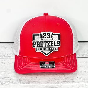 May include: Red and white baseball cap with a white mesh back. The front of the cap features a black and white patch with the text "23 Pretzels Baseball".